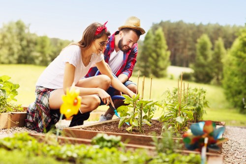 A woman and a man are working together in a well-maintained garden, tending to raised vegetable beds made of wood. The woman is wearing a white t-shirt, plaid shorts, and a pink headband, while the man is dressed in a rolled-up shirt, jeans, and a straw hat. They appear focused on planting or tending to leafy green plants within the garden beds. In the foreground, there are small decorative garden ornaments and a colorful watering can. The background features a lush, green open field with a variety of trees and a bright, sunny sky, suggesting a warm, pleasant day. The garden looks organized with clearly defined borders, soil that appears rich and well-kept, and a combination of flowering plants and vegetables. The outdoor space is spacious, with the natural environment extending into the distance, creating a peaceful and productive gardening scene typical of properties in the Morden area, where gardening and outdoor maintenance services by Lawn Mowing Morden could support such gardens.