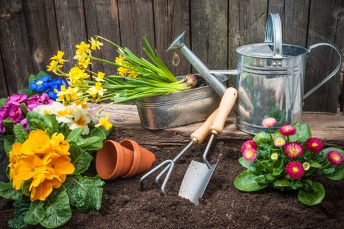 A close-up view of a garden bed in a residential outdoor space, featuring freshly turned dark soil with a variety of potted flowering plants including bright yellow primroses, pink primulas, and purple and blue flowers arranged along a wooden fence. Several gardening tools such as a small hand rake, trowel, and watering can are placed among the plants, indicating recent planting or maintenance activity. The scene suggests early spring gardening work typical of outdoor spaces in Morden, with natural light illuminating the vibrant colors and textures of the flowers, soil, and garden implements, emphasizing a well-maintained and actively cultivated garden area.